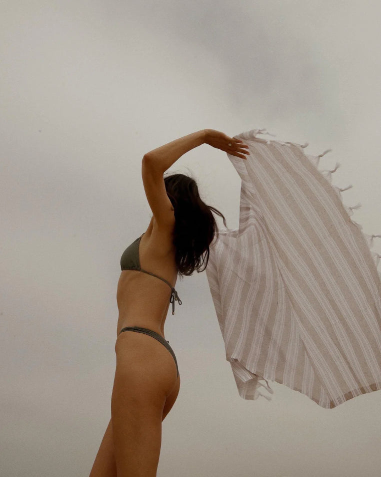 A woman in a dark olive green bikini stands against a cloudy gray sky, holding up a large white and beige vertically striped beach towel with tassels.