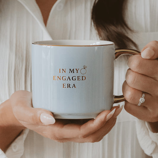 Sweet Water Decor In My Engaged Era white coffee mug with gold wording held by woman in cream knit top